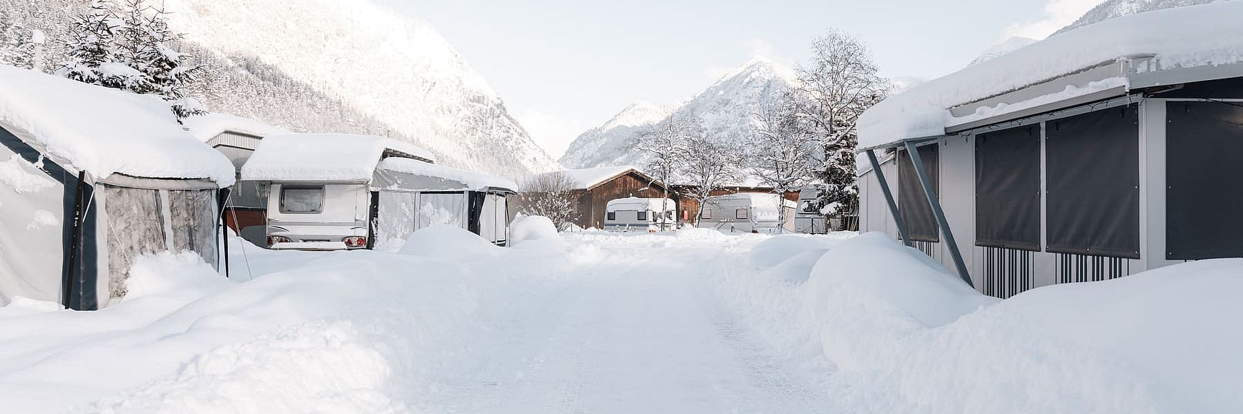 Winterlicher Campingplatz mit tief verschneiter Zufahrt, Wohnwagen und Alpenpanorama im Hintergrund