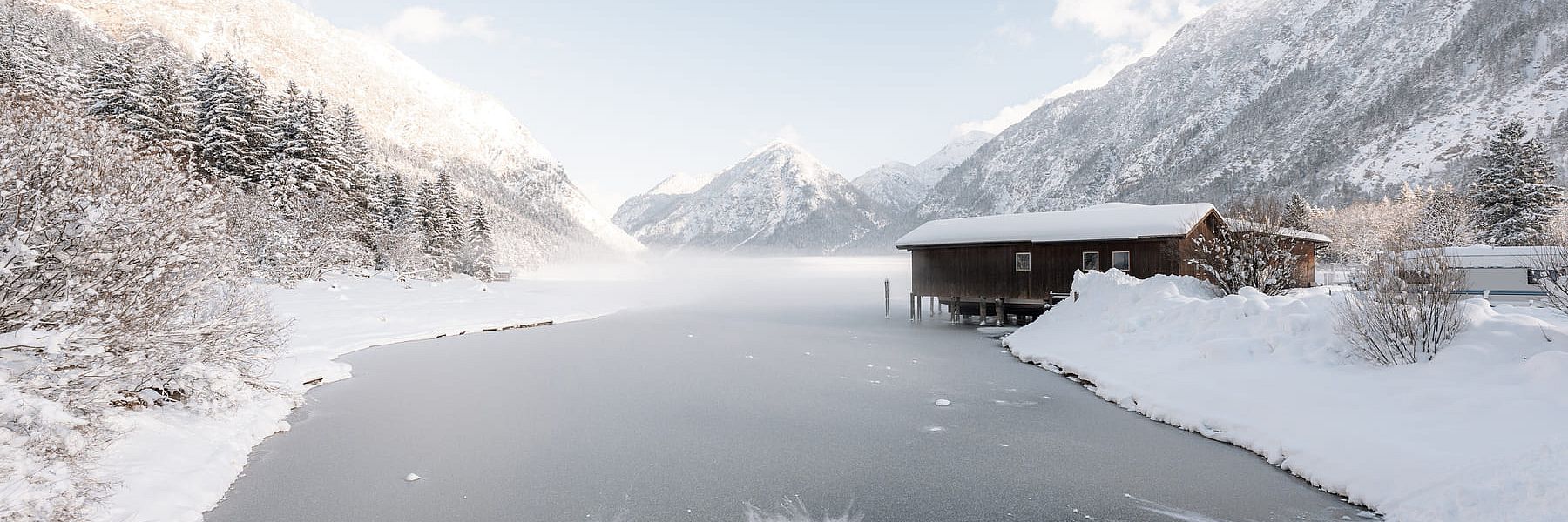 Teilweise zugefrorener See mit Holzhaus am Ufer, umgeben von schneebedeckten Bergen im Winter